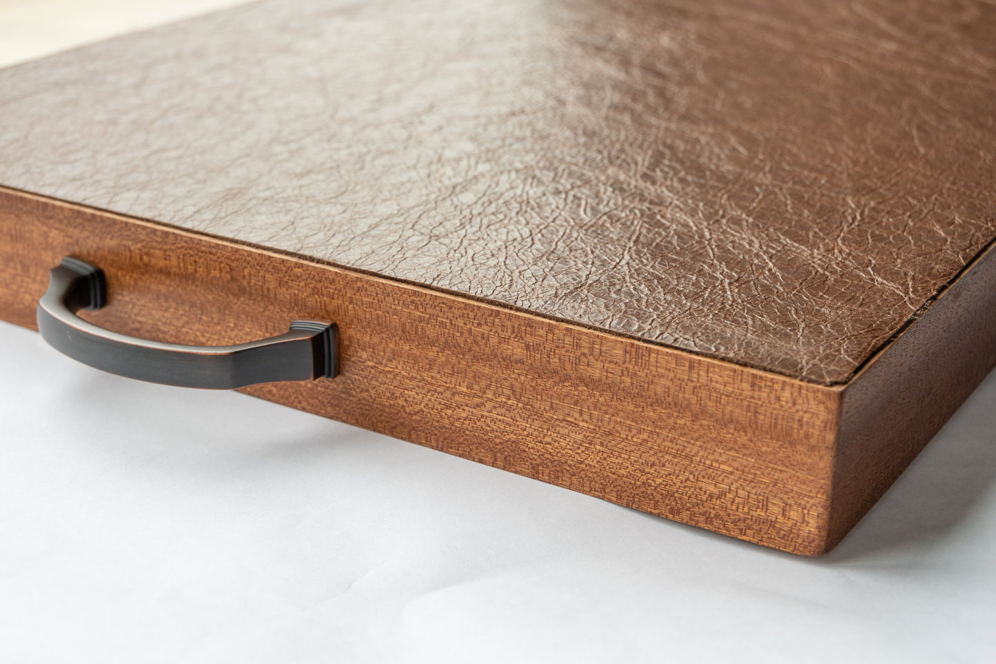 A close-up of the leather backing of our luxury Serving Tray. The thick, soft, and supple leather is attached to the back of solid oak wood and  meets the edge of the mahogany lip that features the wood's natural grain in stunning detail. A black handle protrudes from the side. 