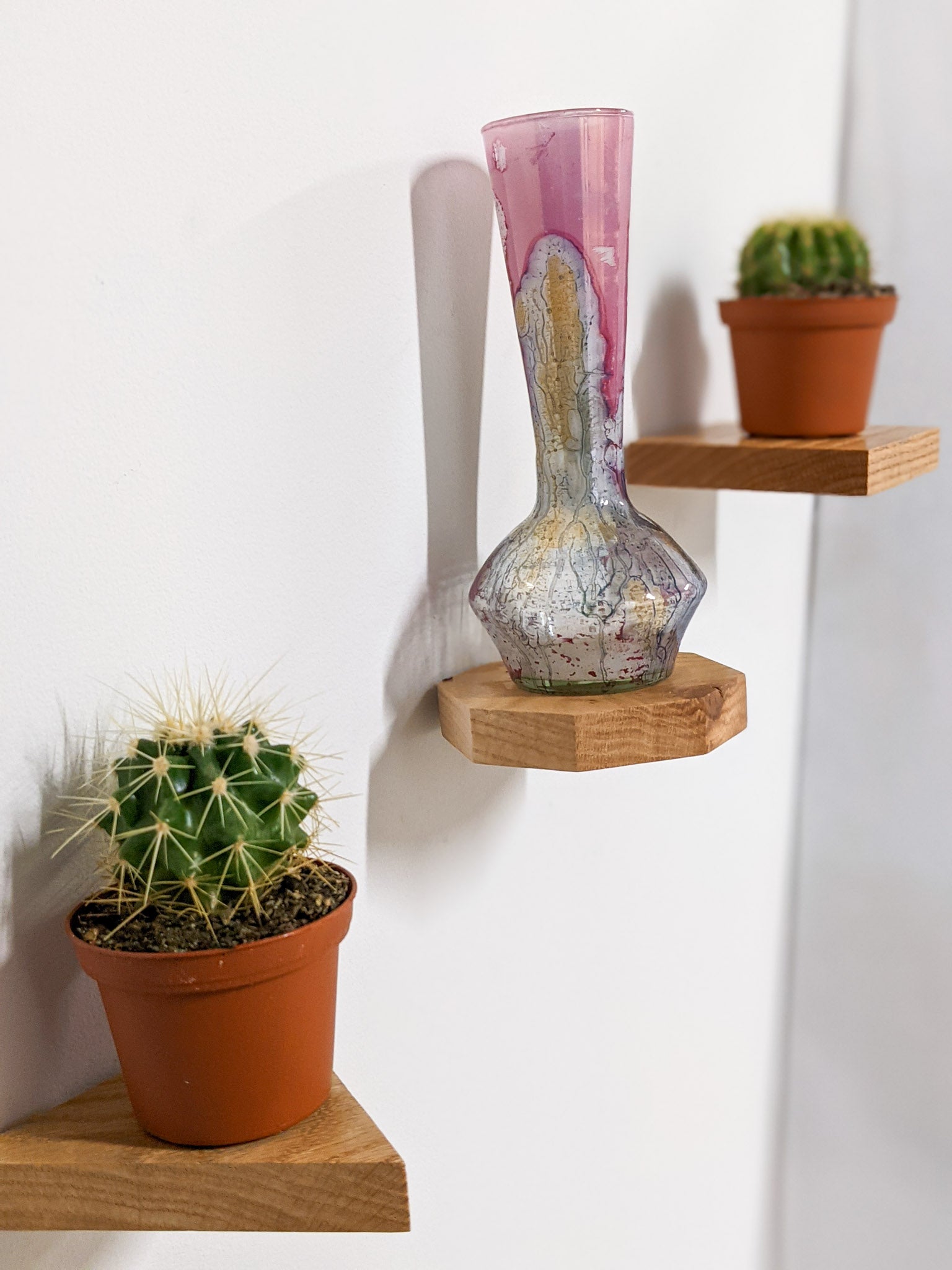 Three mini oak shelves ascend a wall. The first is a triangle oak floating shelf, the second - an octagon oak floating shelf that holds a vase, and the third is a square oak floating shelf that holds a cactus.