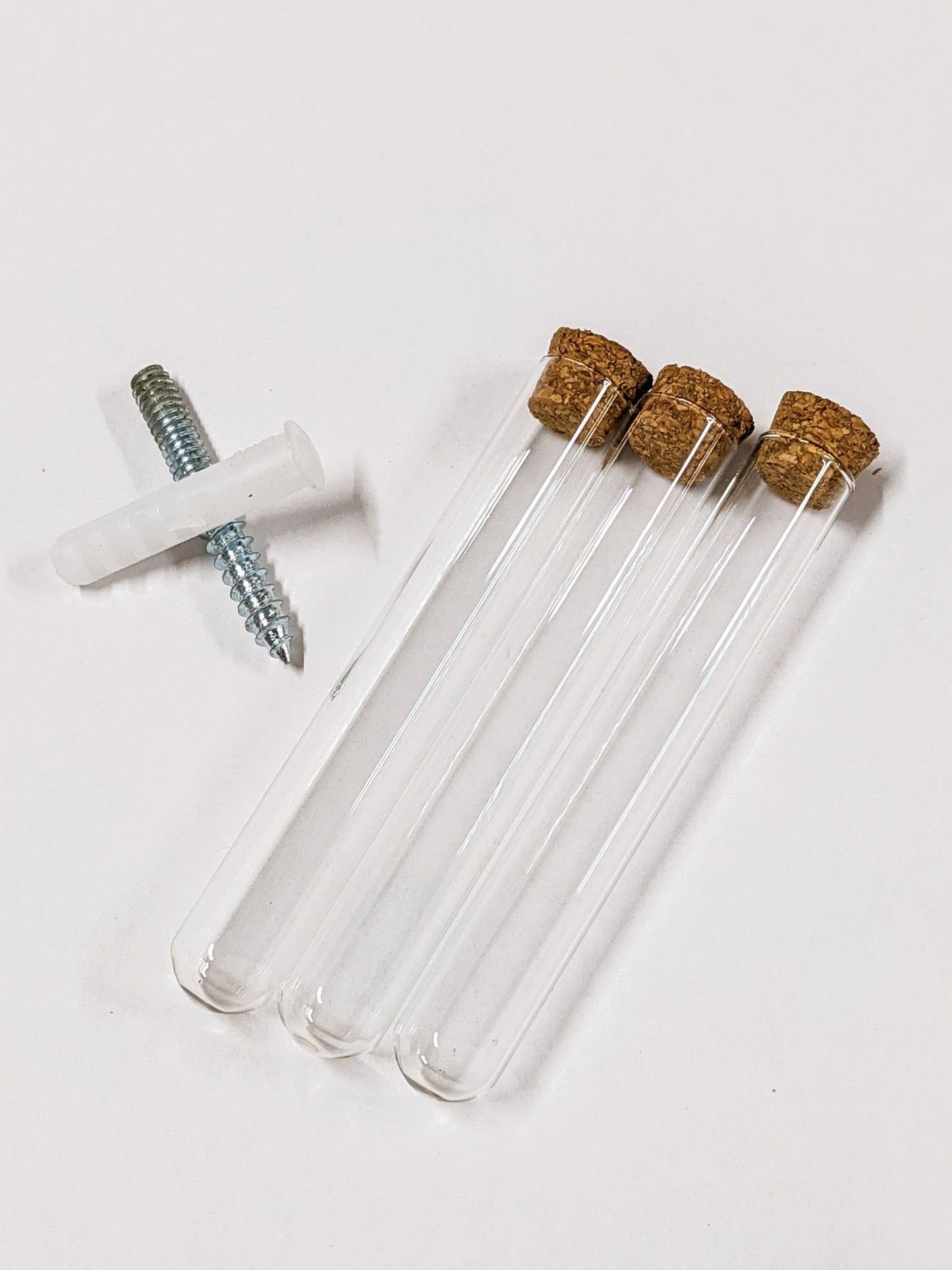 Three propagation station test tubes lay next to each other on a white background. A silver screw and white anchor are placed in an X pattern with their centers touching. The test tubes are corked.