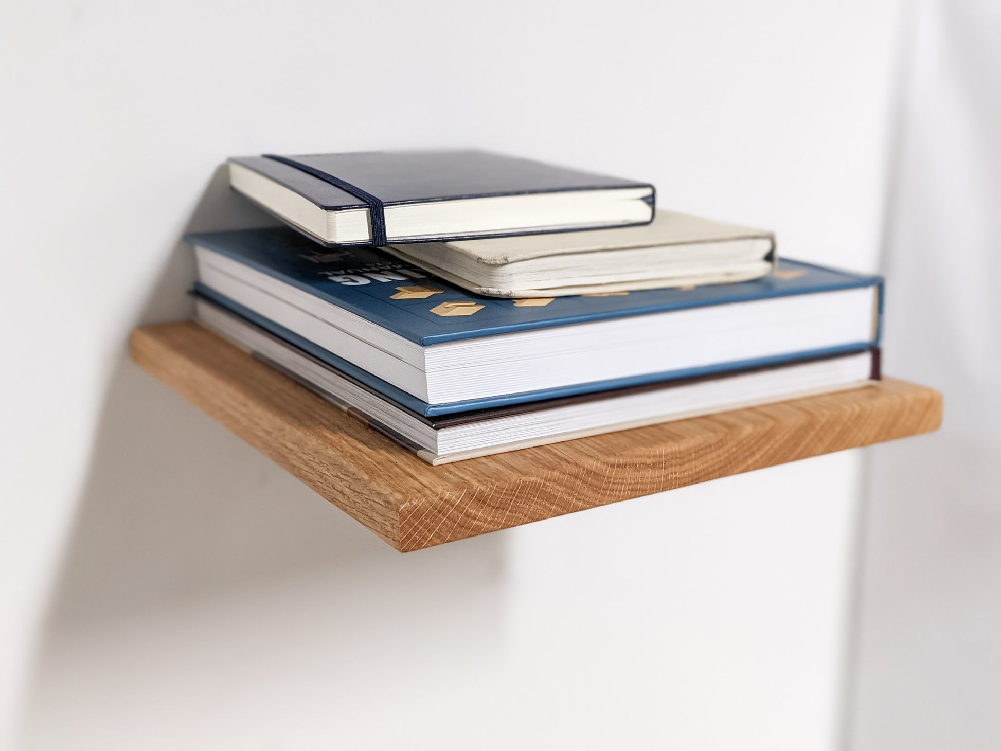 A customized medium oak floating shelf holds a stack of four books in blue and gray shades.