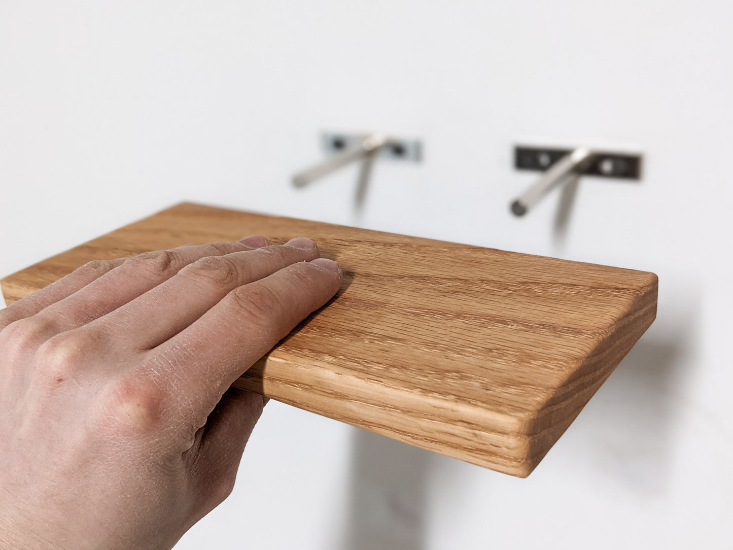 A hand holds a small, rectangular, floating oak wood shelf in the foreground. In the background, two silver rods protrude from the invisible bracket.