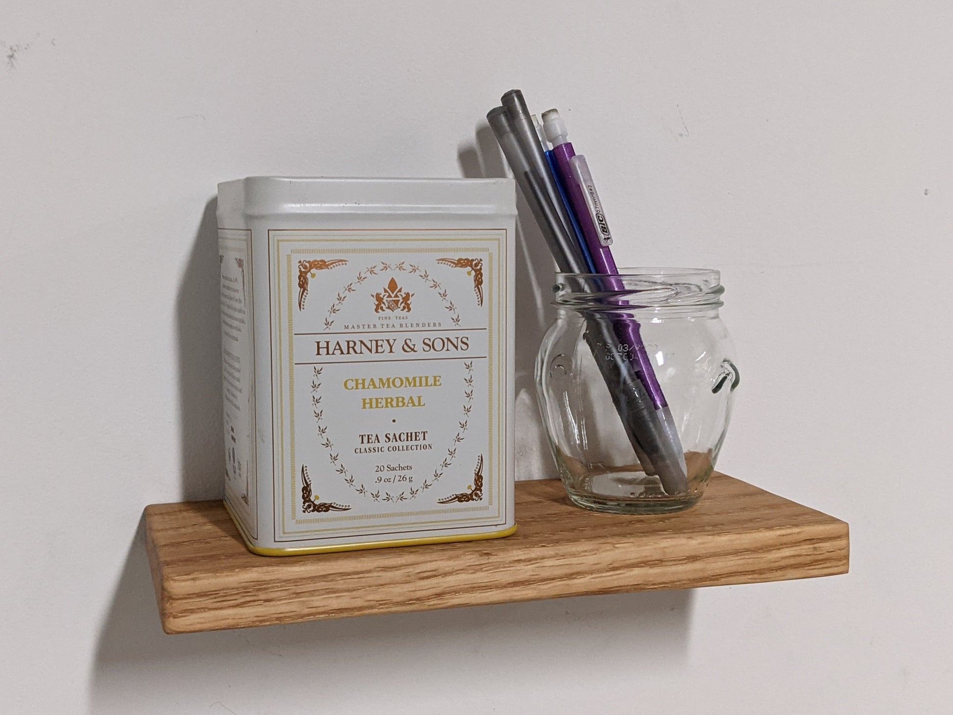 A minimalist oak floating shelf is easily mounted onto a wall and displays a white Harney and Sons, Chamomile Herbal tea bin and a clear round pencil holder. There are two pens and two pencils in the pencil holder.