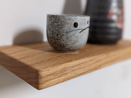 A close-up on a custom oak floating shelf with soft rounded corners and smooth natural surface. A grey, Japanese style tea mug, sits atop the floating shelf. 