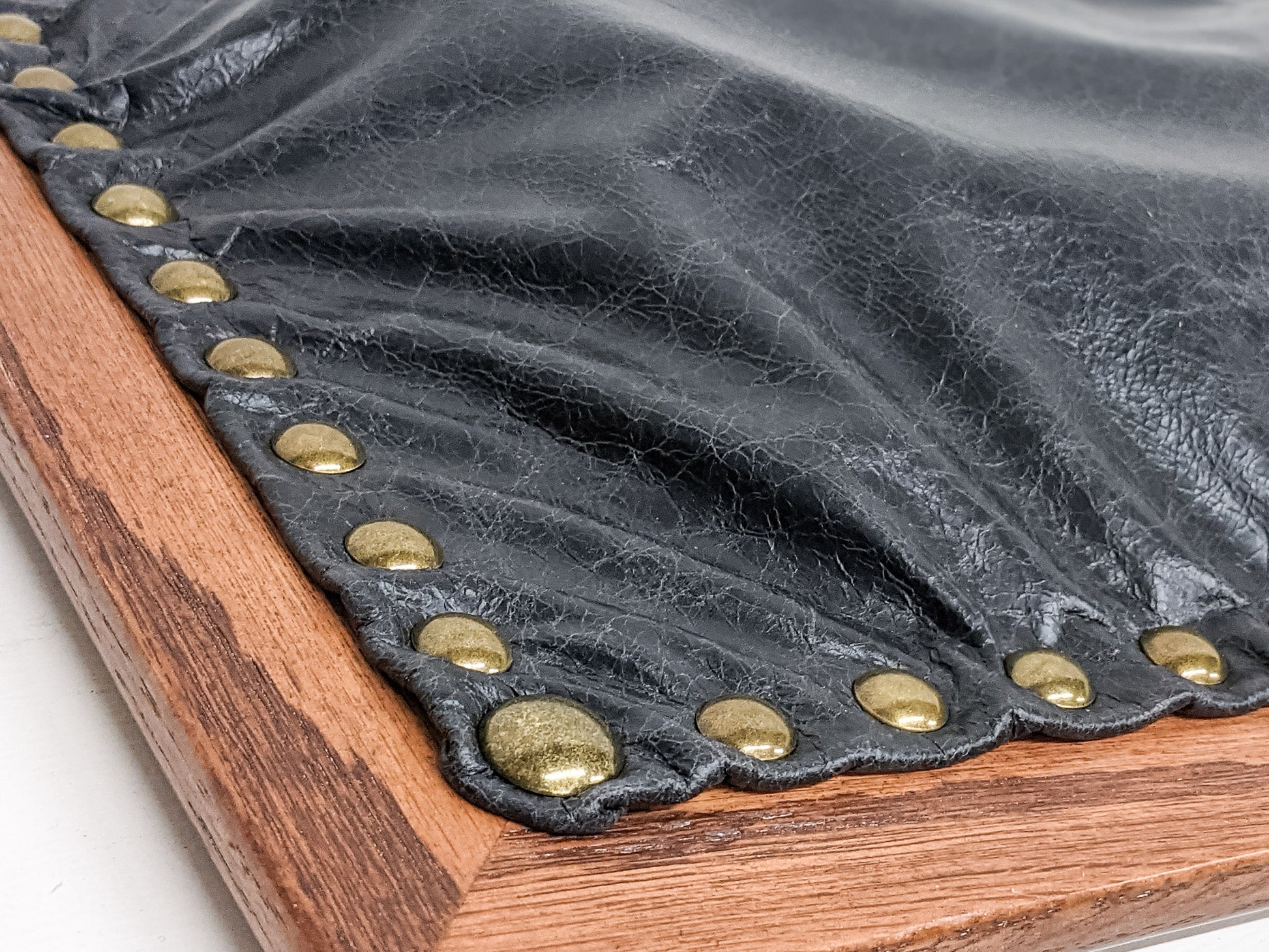 A close-up of our black leather option for our Wooden Writing Lap Desk. The corner of the mahogany lap desk is in view and the textured black leather grain is attached with a number of round and gleaming brass pins. 