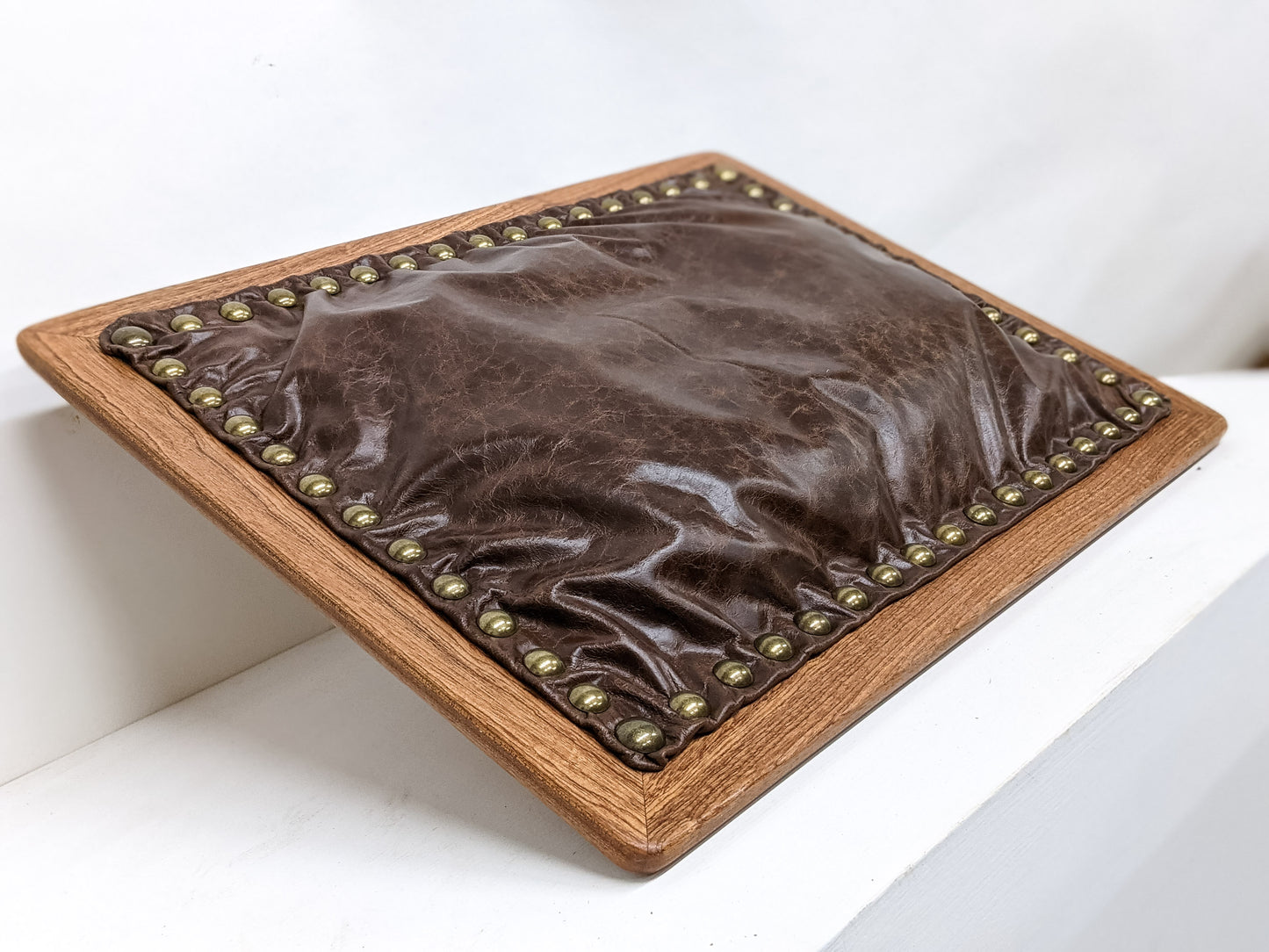 The bottom of our Leather Cushion Writing Lap Desk is shown. The mahogany wood frames a bulging bean filled, dark brown, leather cushion that is securely attached by 56 shining brass pins.