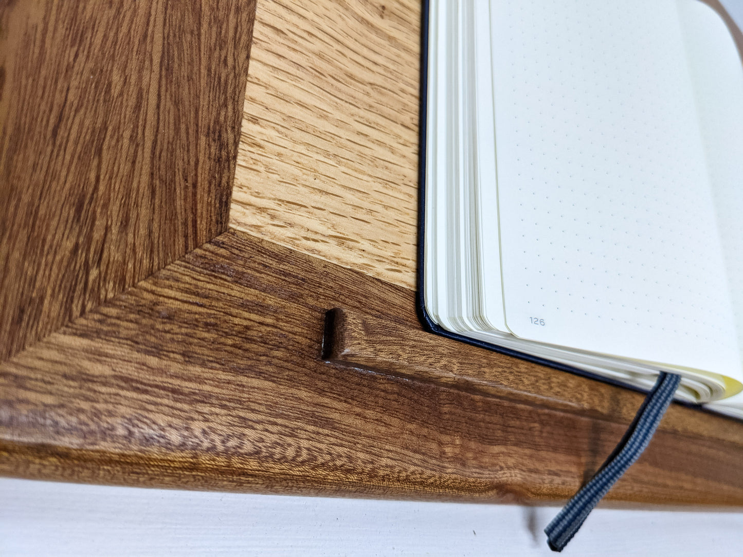 A close-up of the protruding mahogany lip that serves as a book rest on our wooden writing lap desk in oak and mahogany. A mahogany wood frame meets an oak interior to create an understated and elegant meeting of the woods. A bullet journal is opened to page 126 and is blank. A navy fabric book marker saves a spot in the journal. 