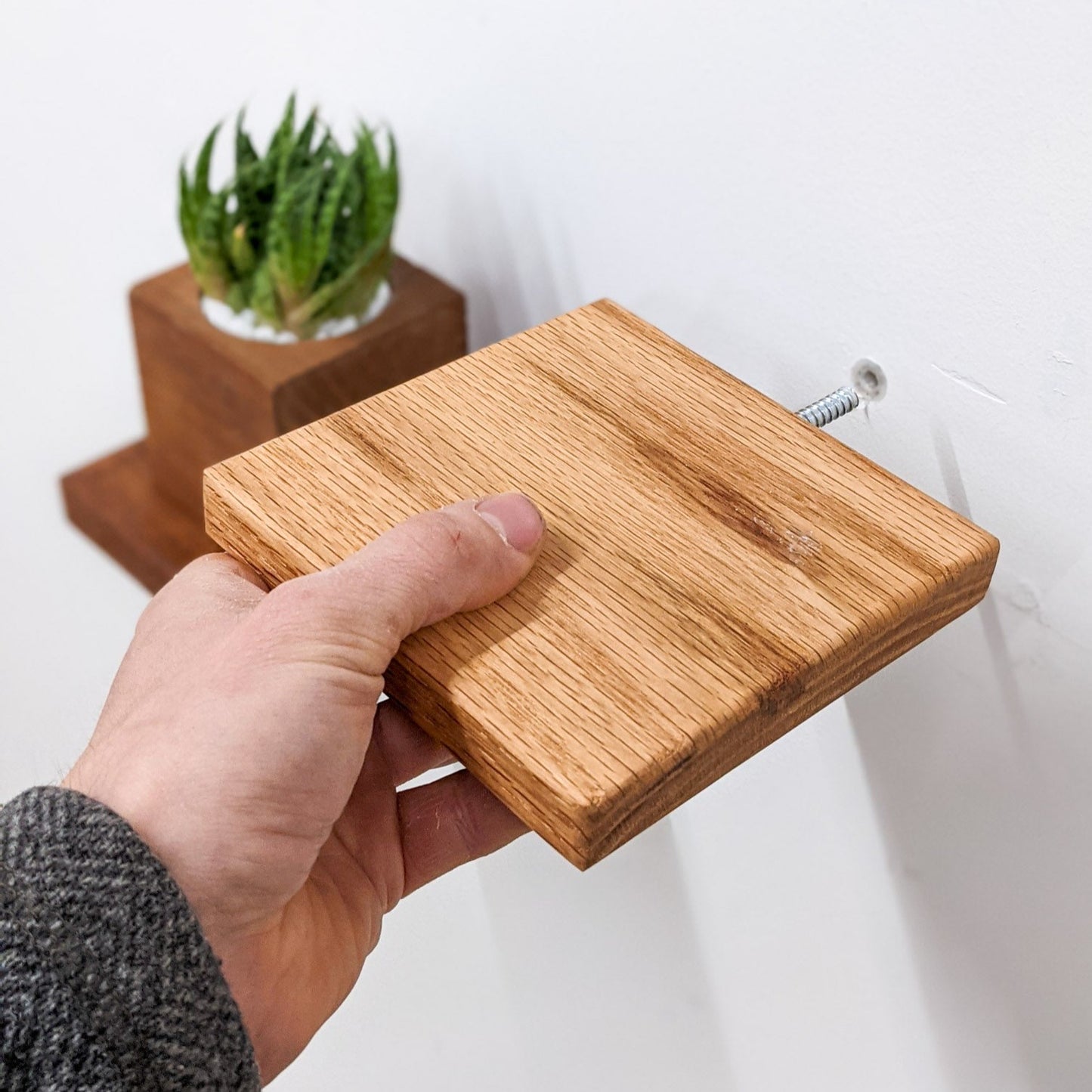 A hand installs a small wooden oak square shelf. In the background, an out of focus mahogany square wooden shelf is already installed and holds a square planter with a succulent.