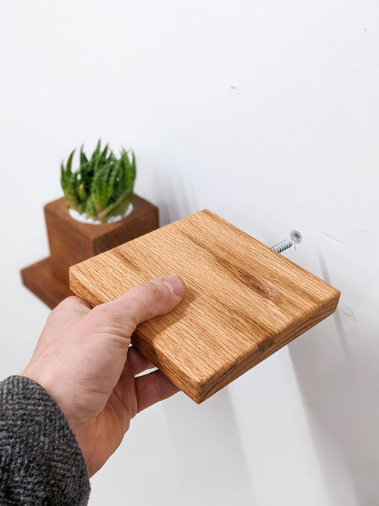 A hand installs a small wooden oak square shelf. In the background, an out of focus mahogany square wooden shelf is already installed and holds a square planter with a succulent.