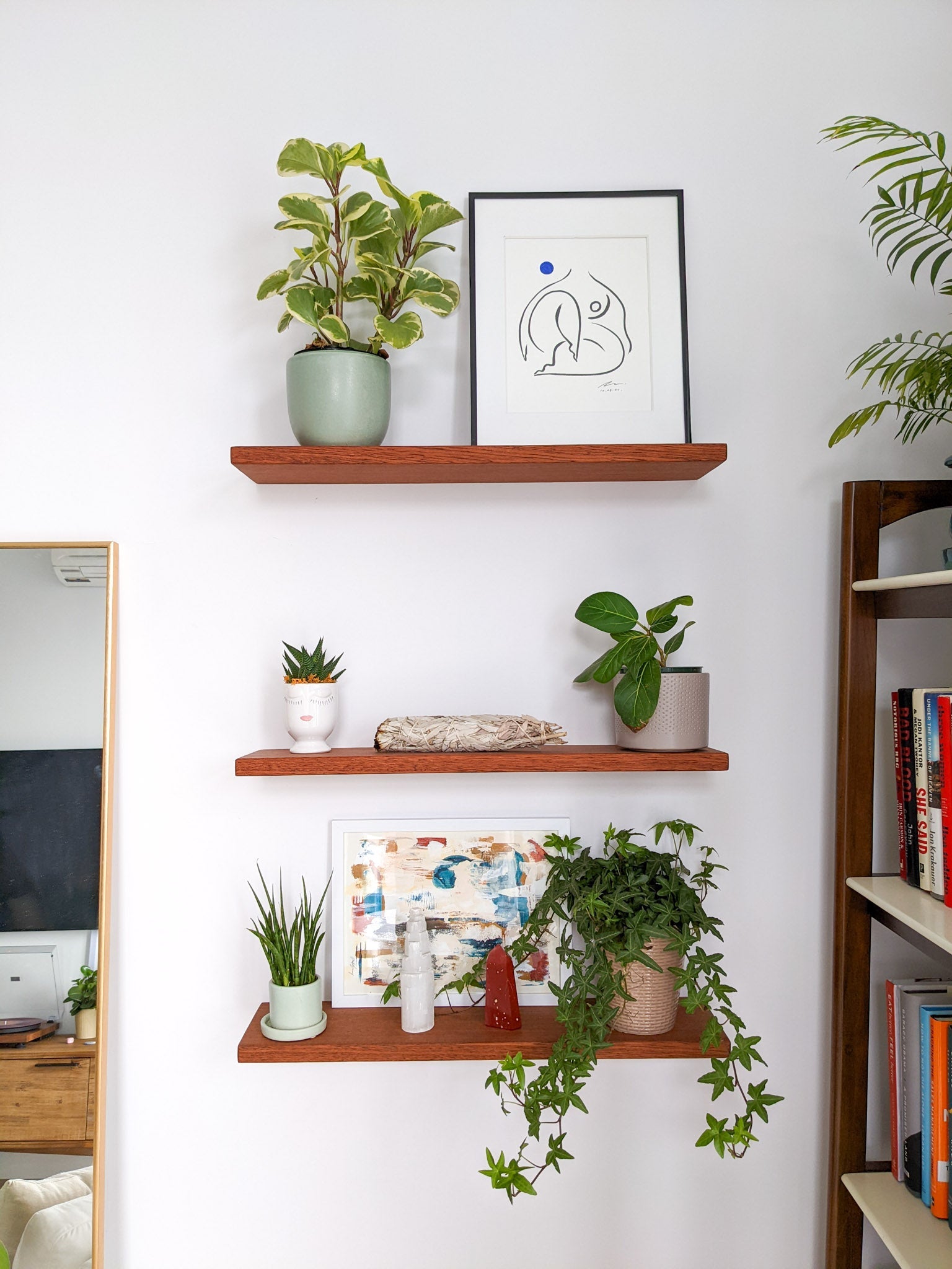 Three medium sized mahogany floating shelves are stacked on top of the other. The highest shelf holds a rubber plant in a blue planter and an impressionist painting; the middle shelf holds a succulent in a novelty face planter, a white sage stick, and a small ficus in a gray pot; and the lowest shelf holds a fern and pickle plant and two crystal stalagmites.