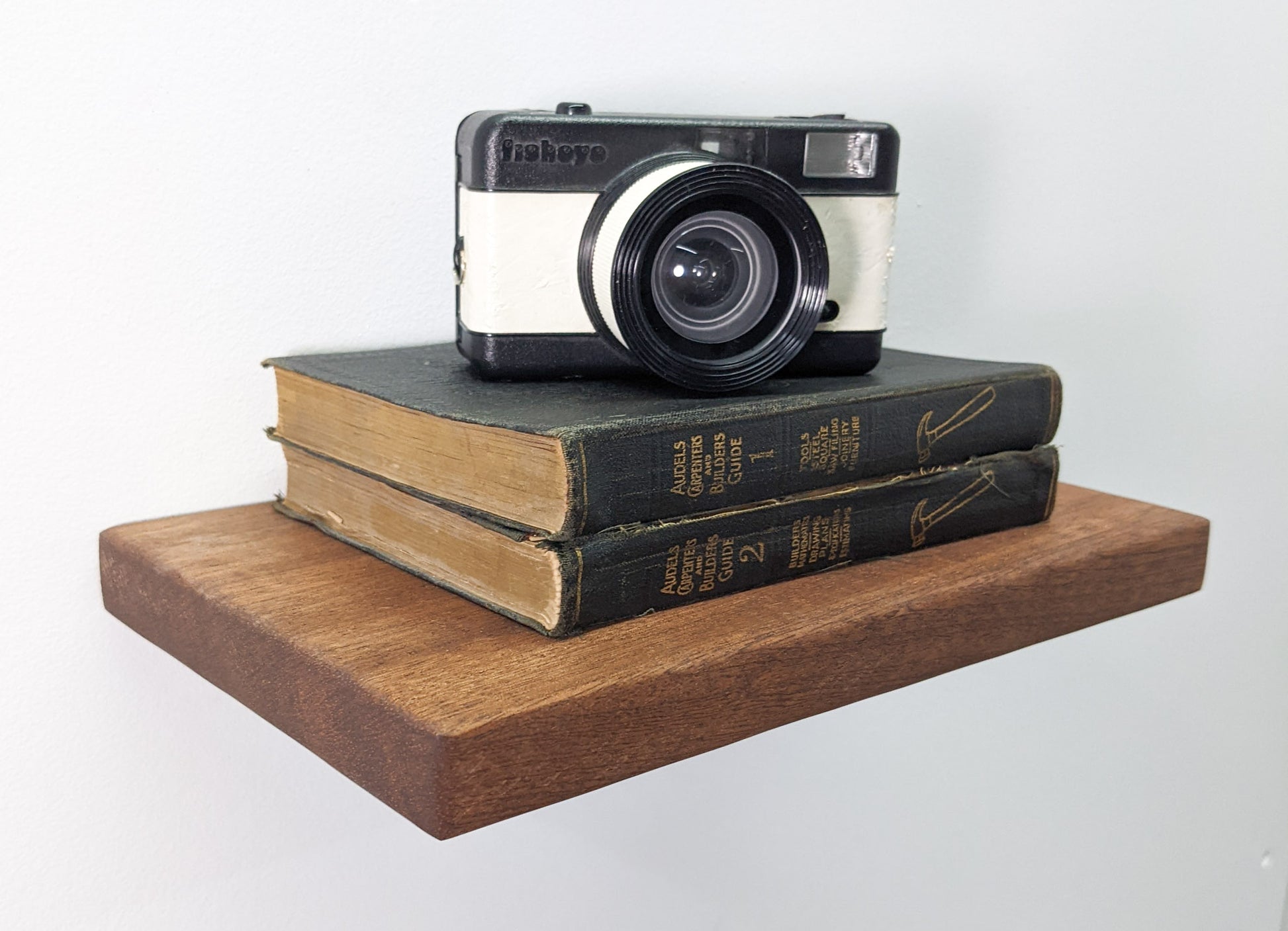 A side view of our medium mahogany floating shelf with bevelled soft edges. It holds holds two black leather bound books with gold embossed writing, reading, "Audells Carpenters and Builders Guide" Atop the books, an old-fashioned camera sits.