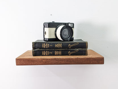 A sturdy solid wood mahogany floating shelf holds two black leather bound books with gold embossed writing, reading, "Audells Carpenters and Builders Guide" atop the books, an old fashioned camera sits.