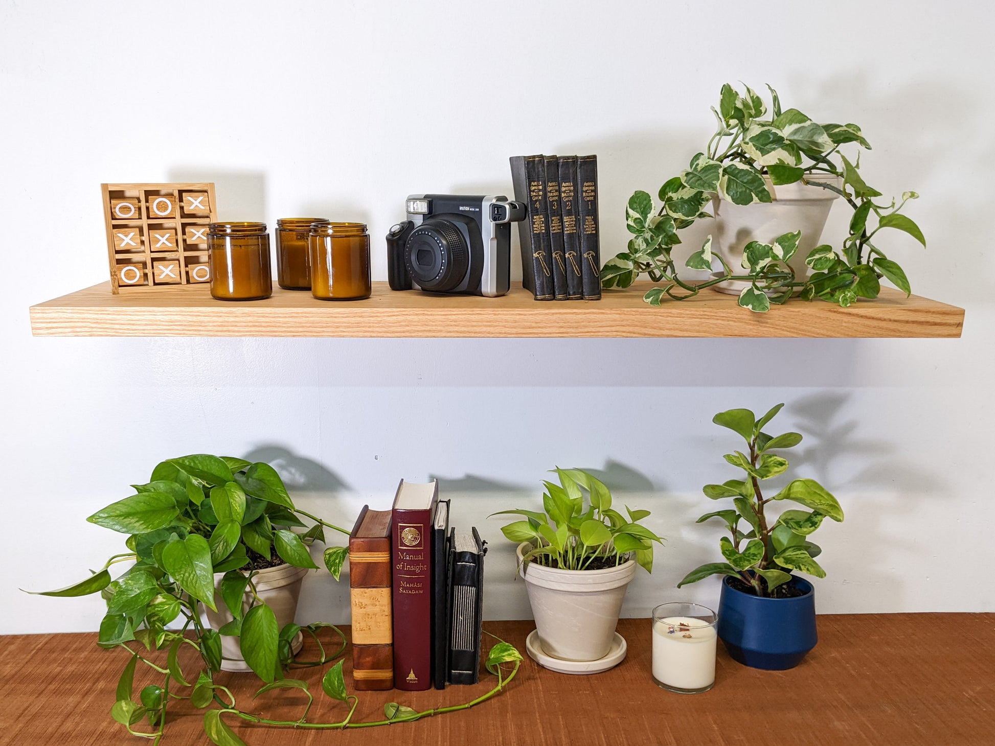 The front view of a medium oak floating shelf. On top, four black leather bound books are displayed, next to three vintage candles, and a birds nest fern that sits within a wood round planter.