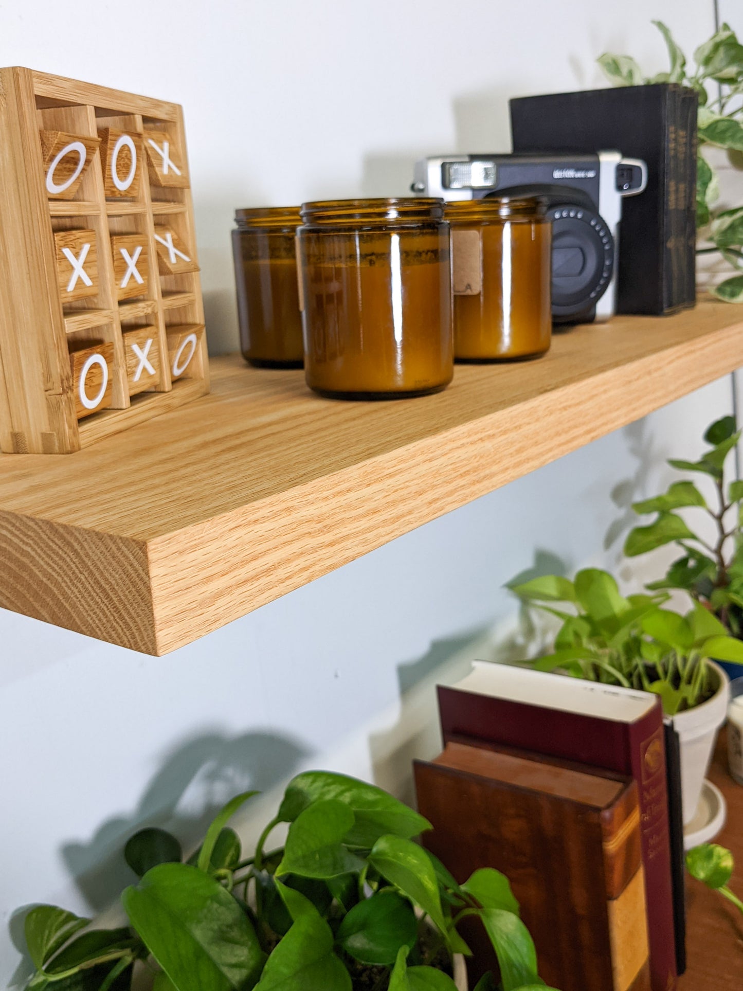 The side view of a long oak floating shelf that displays three amber candles, a decorative wooden tic-tac-toe game, three vintage books, and a vintage camera.