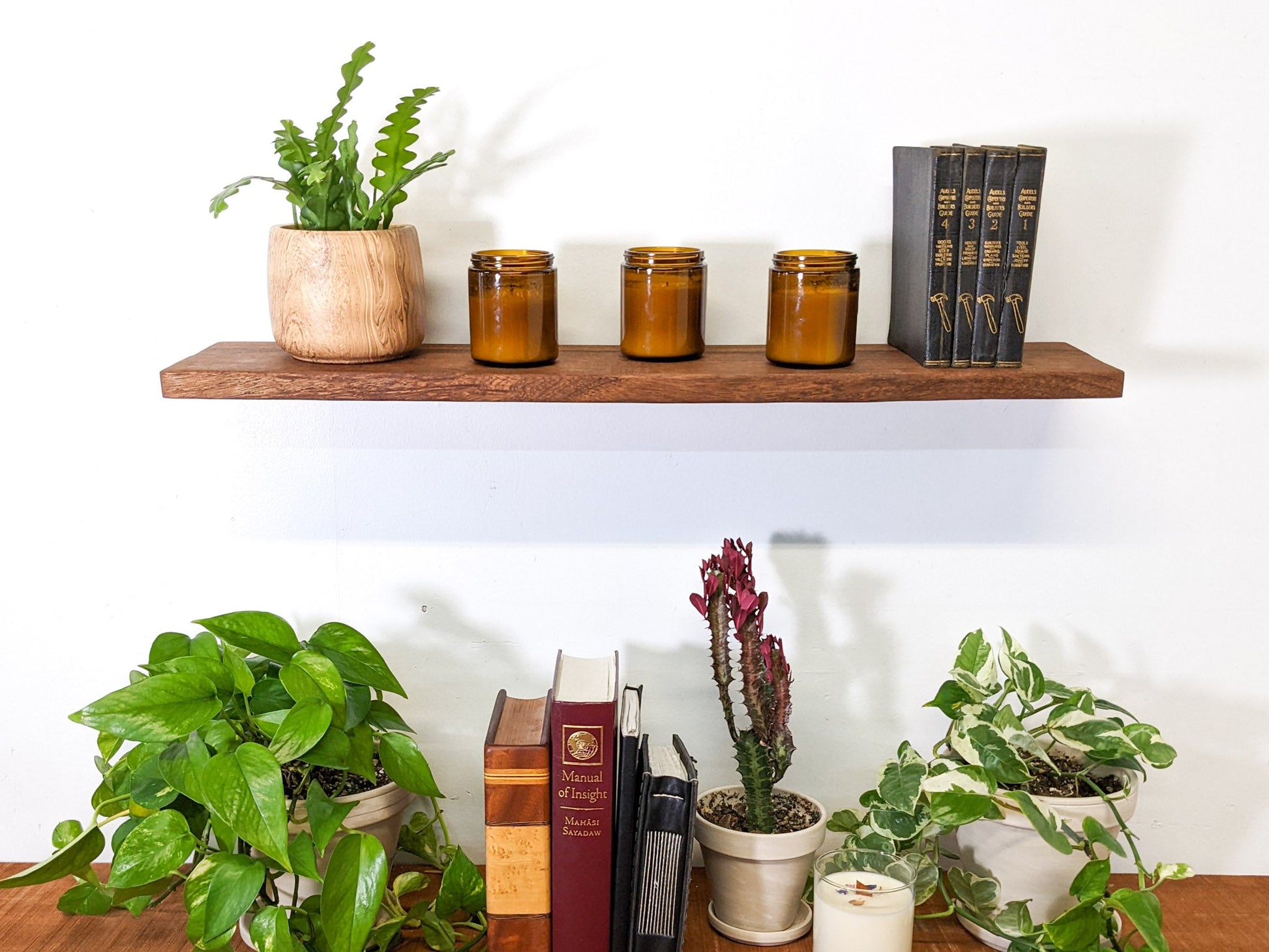 The front view of a medium mahogany floating shelf. On top, four black leather bound books are displayed, next to three vintage candles, and a birds nest fern that sits within a wood round planter.