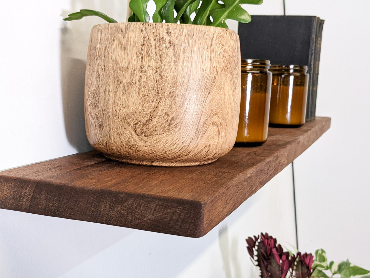 A close-up of the soft bevelled edges of a medium-duty mahogany floating shelf, a crispy fern sits on top with two vintage candles beside it.