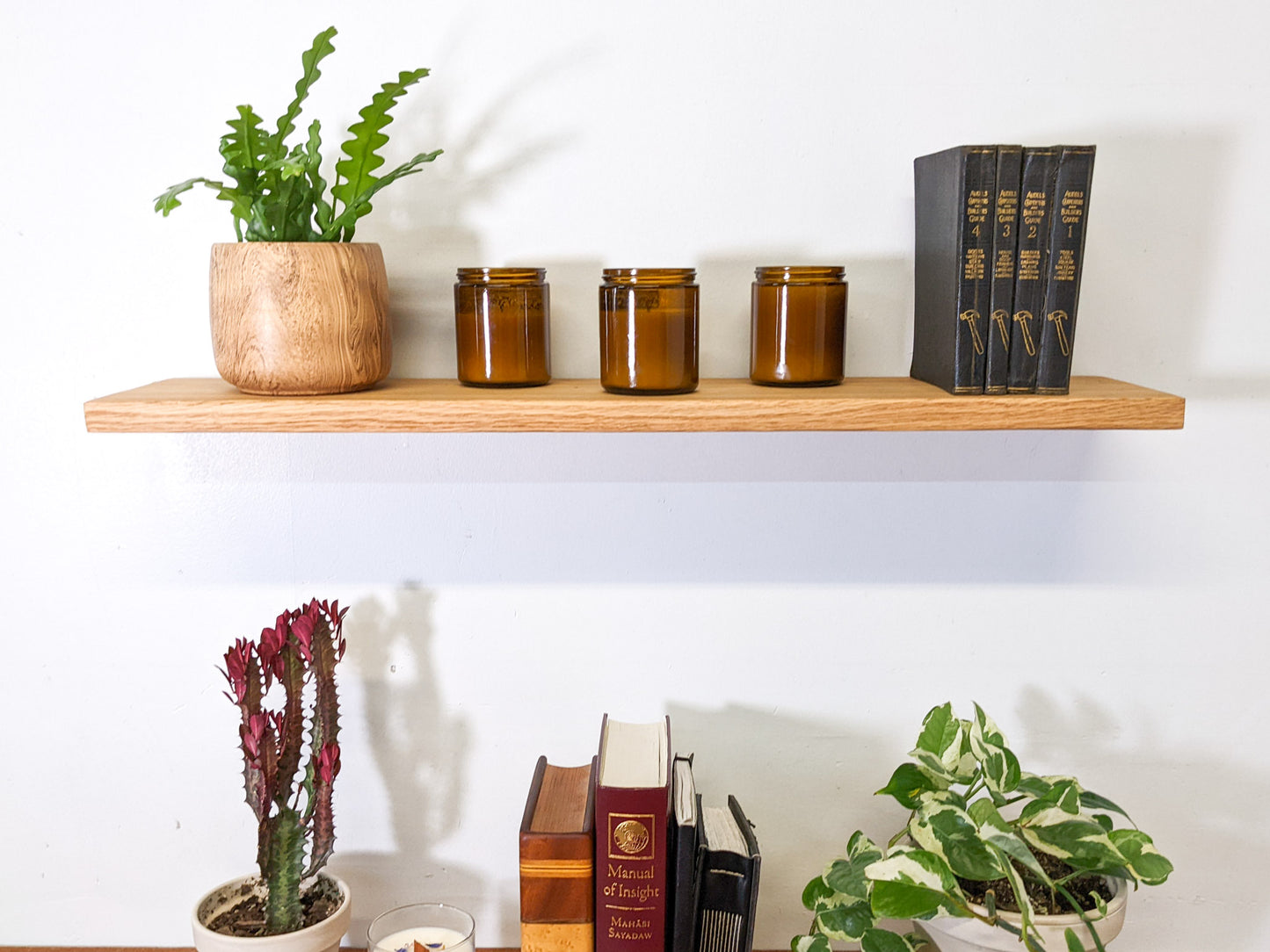 The front view of a medium oak floating shelf. On top, four black leather bound books are displayed, next to three vintage candles, and a birds nest fern that sits within a wood round planter.