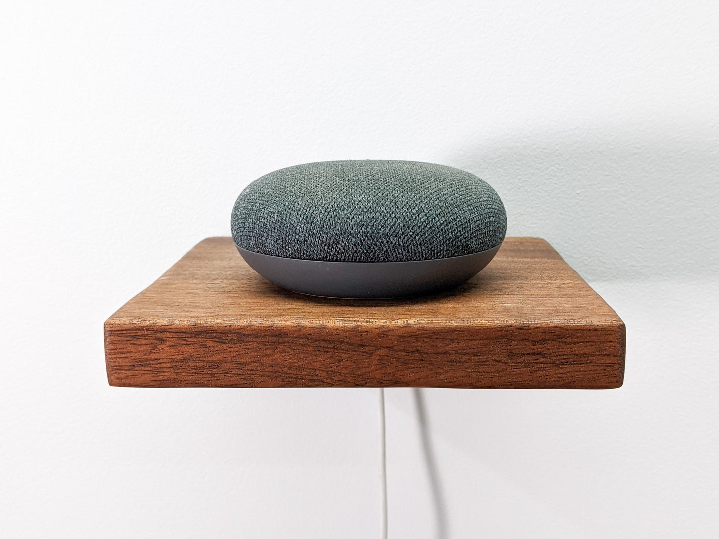 A front-on view of the mahogany cord management shelf. The mahogany shelf has clean lines and beveled edges that create a minimalist design that pops against the white wall. A small round speaker sits on top and a white cord is threaded through a drilled hole in the shelf.