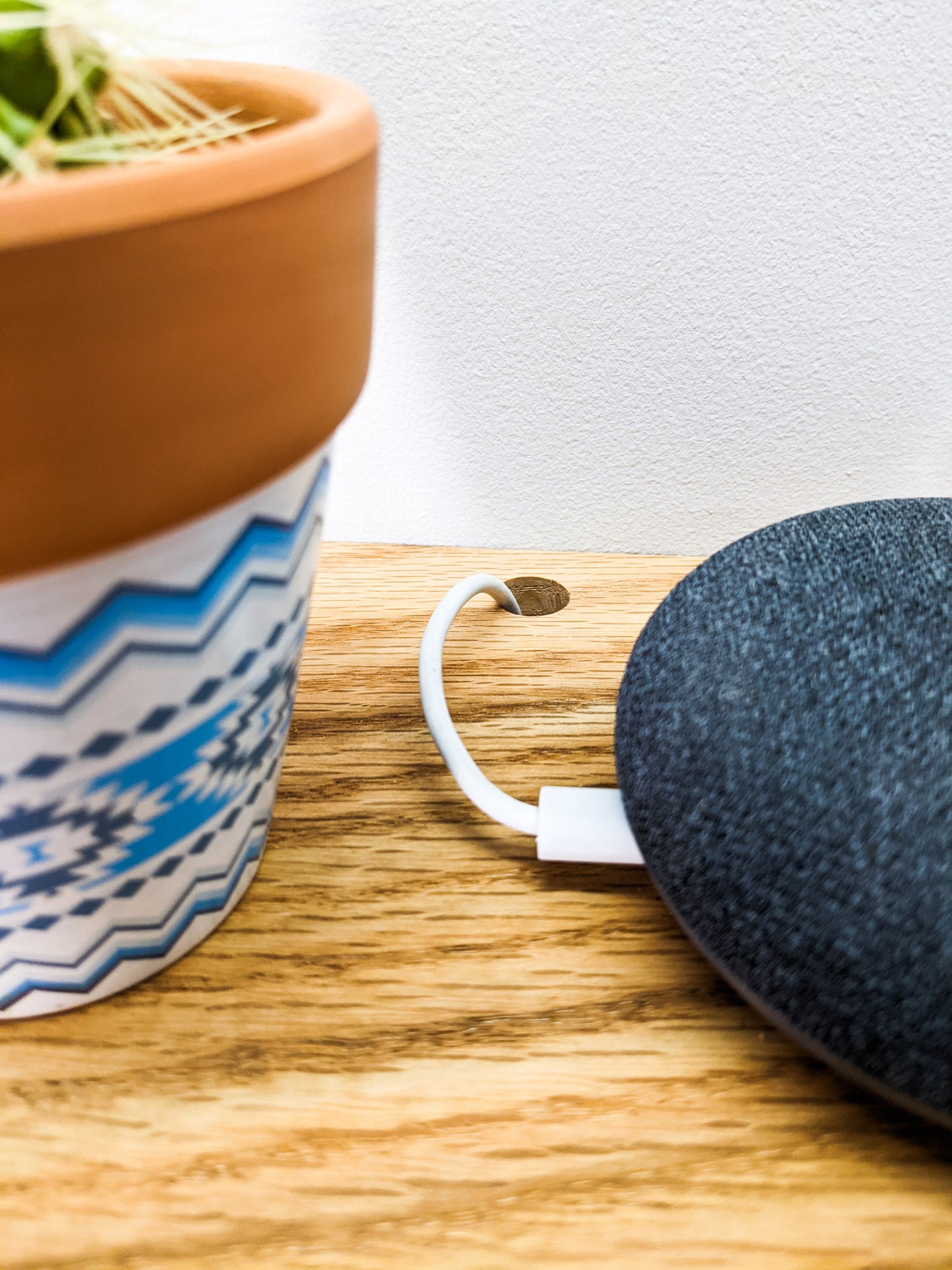 From above we see NookWoodworking's rectangular oak floating shelf with a hole for cable management. A round speaker sits on top and a white cord is threaded through a small, cleanly drilled hole in the back of the shelf. To the left, a white and blue unique planter sits. 