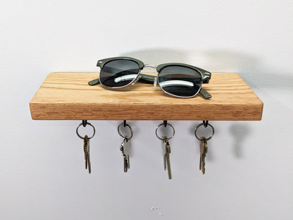 A head-on view of NookWoodworking's oak key hook shelf. The soft fine grain of the oak wood displays natural whirls on the wall-mounted base. A pair of black sunglasses rest on top. Below, four hanging black key hooks hold eight keys. Four keys are bronze and four are silver.