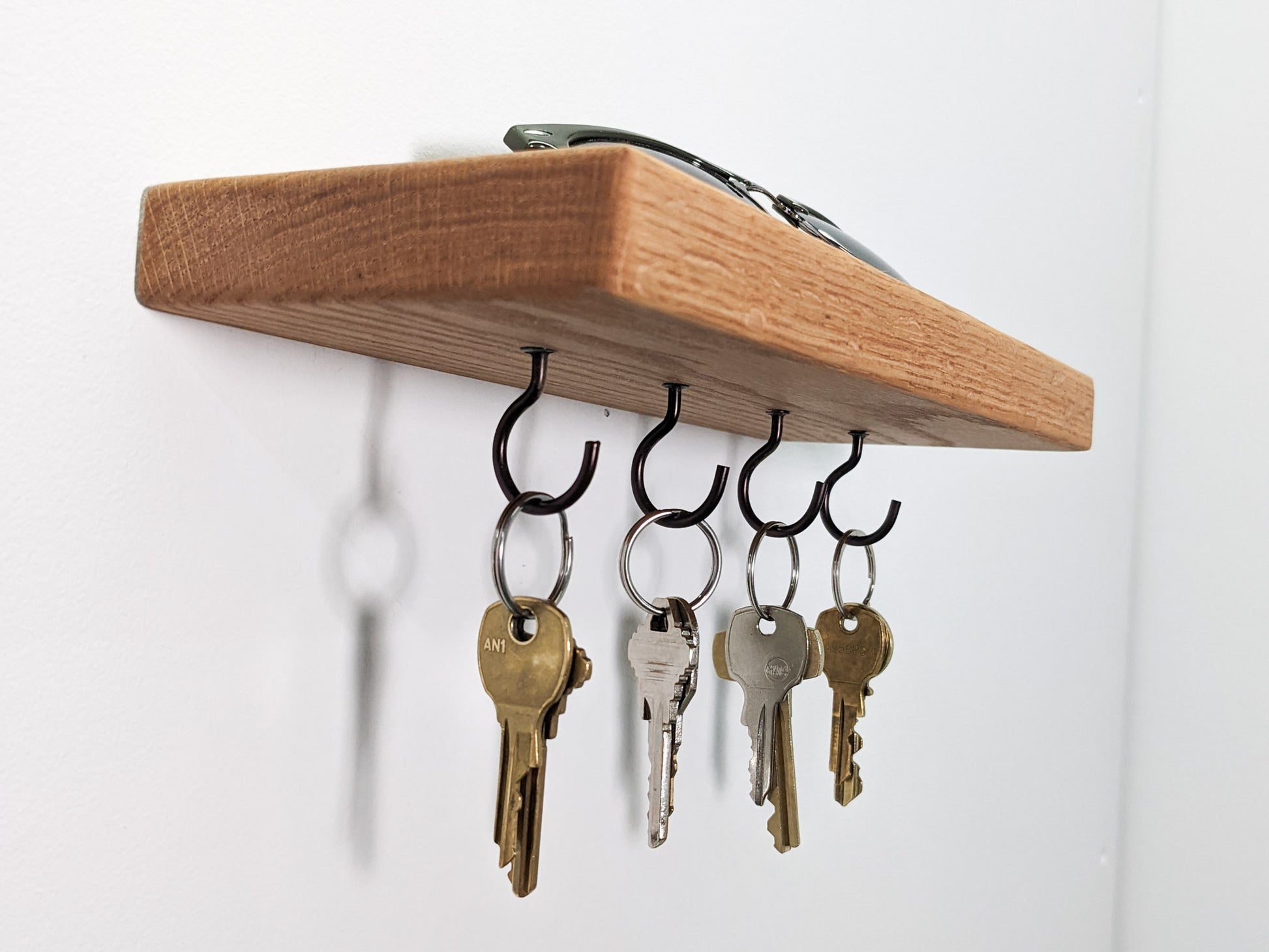 Four black key hooks hang securely from a wall-mounted floating oak shelf . Four keys are bronze, four are silver. The keys cast a slight shadow onto the white wall behind them. On top of the shelf sits a pair of sunglasses. 