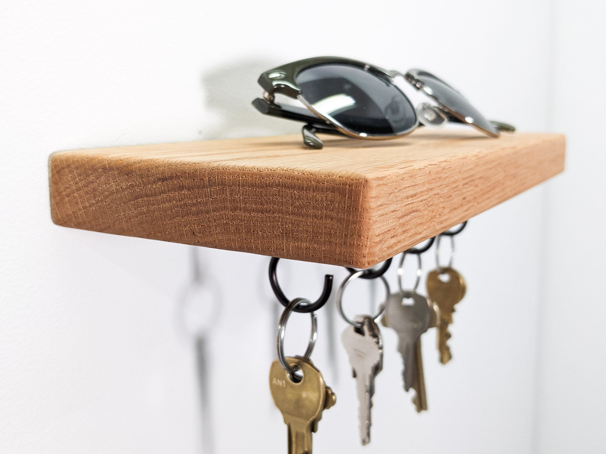 A side view of a floating key hook shelf. The natural grain of the light oak wood presents as vertical-scratch-like-marks that reach up and over the soft beveled edges of the corners. On the secure oak base sits a pair of black sunglasses. Below, four hanging black key hooks hold eight keys. Four keys are bronze and four are silver.