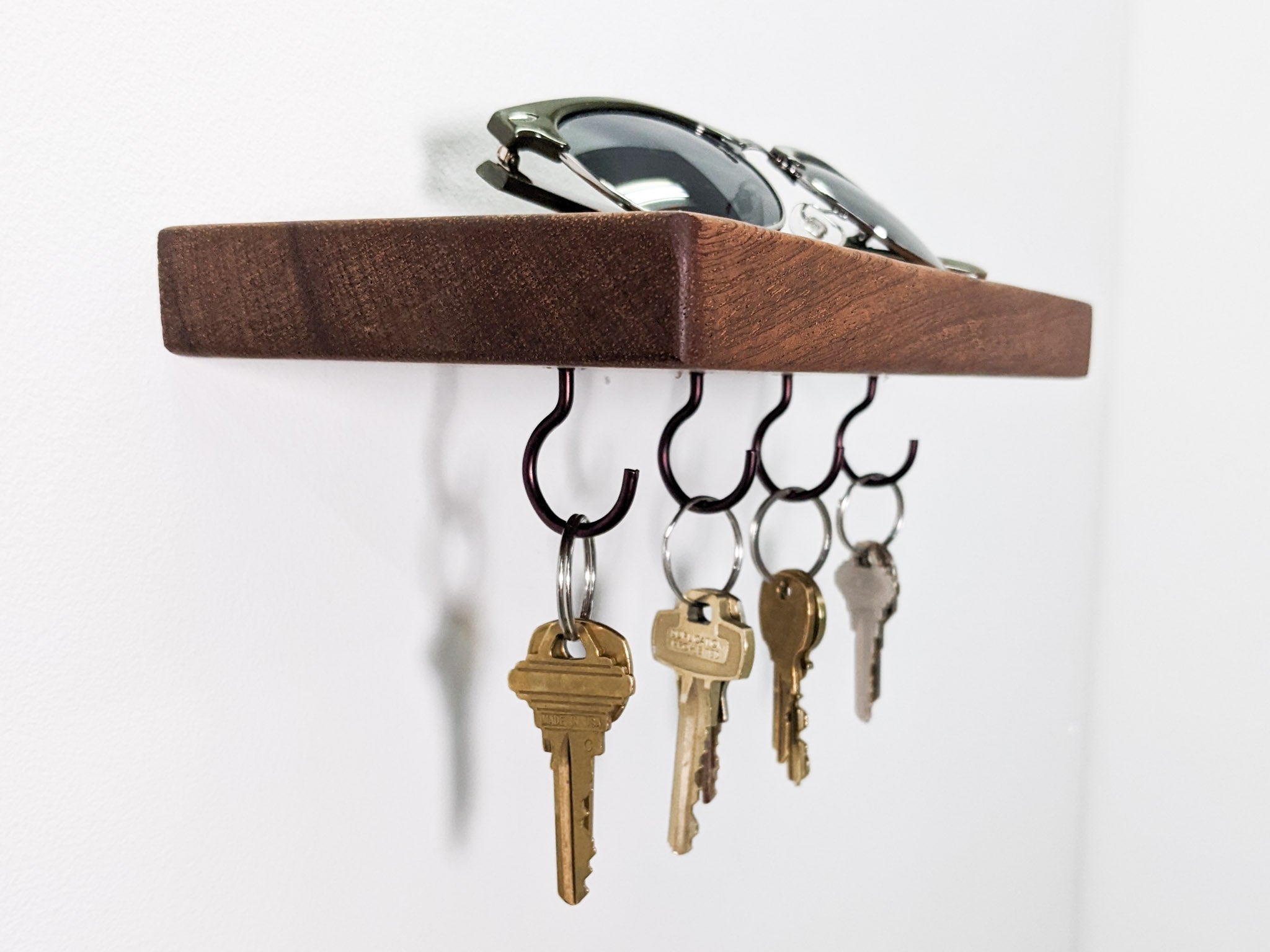 Four black key hooks hang securely from a wall-mounted floating mahogany shelf . Four keys are bronze, four are silver. The keys cast a slight shadow onto the white wall behind them. On top of the shelf sits a pair of sunglasses.