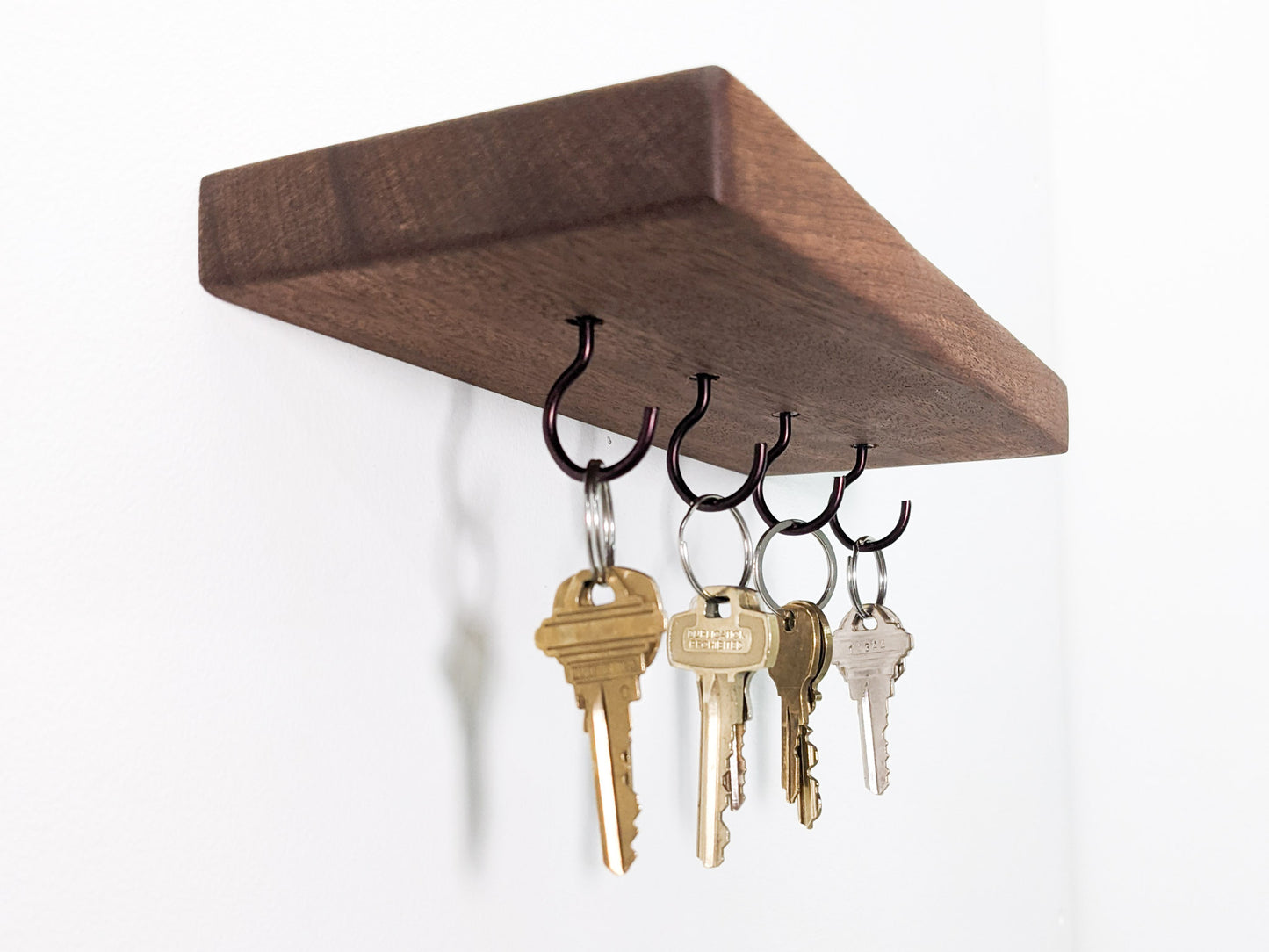 A side view of a floating key hook shelf. The natural grain of the brown mahogany wood presents as vertical-scratch-like-marks that reach up and over the soft beveled edges of the corners. On the secure mahogany base sits a pair of black sunglasses. Below, four hanging black  key hooks hold  eight keys. Four keys are bronze and four are silver. 