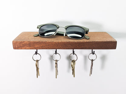 A head-on view of NookWoodworking's mahogany key hook shelf. The soft fine grain of the brown mahogany wood displays natural whirls on the wall-mounted base. A pair of black sunglasses rest on top. Below, four hanging black key hooks hold eight keys. Four keys are bronze and four are silver.