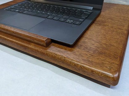 The Leather Cushion Lap Desk is angled towards the camera. The wood mahogany of the desk gleams while the soft rounded edges create a pleasing look. A black laptop is opened on the desk.