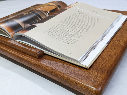 The Leather Cushion Lap Desk is angled towards the camera. The wood mahogany of the desk gleams while the soft rounded edges create a pleasing look. A book is opened on the desk and rests on the book lip. The page reads, "Boxes & Chests".