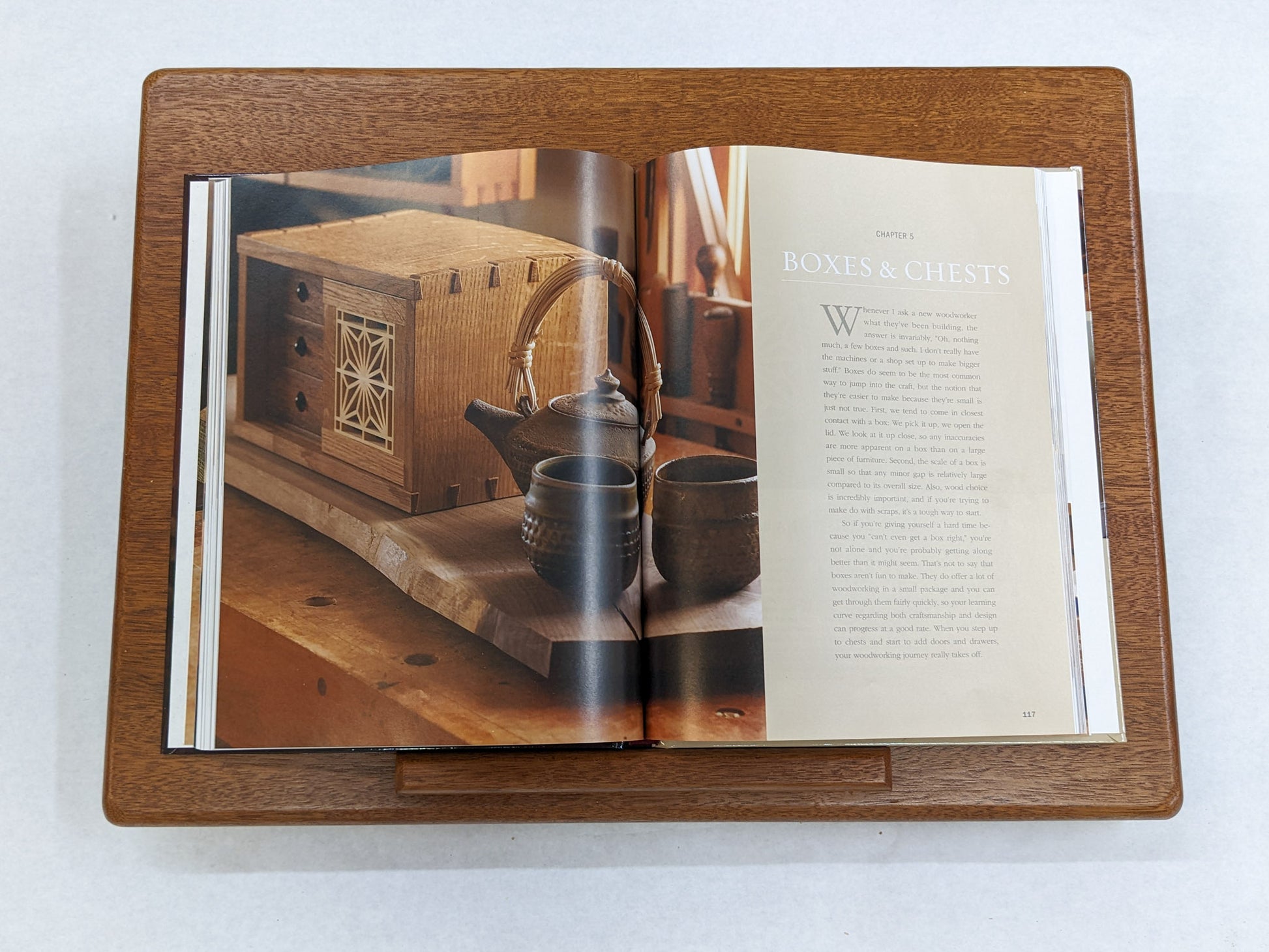 The Leather Cushion Lap Desk is viewed from above. A large book is resting on the protruding mahogany lip of the desk. The book is opened to a page that has a photo of a box, tea kettle, and two tea mugs, on the right page it reads, "Boxes & Chests" in white letters.