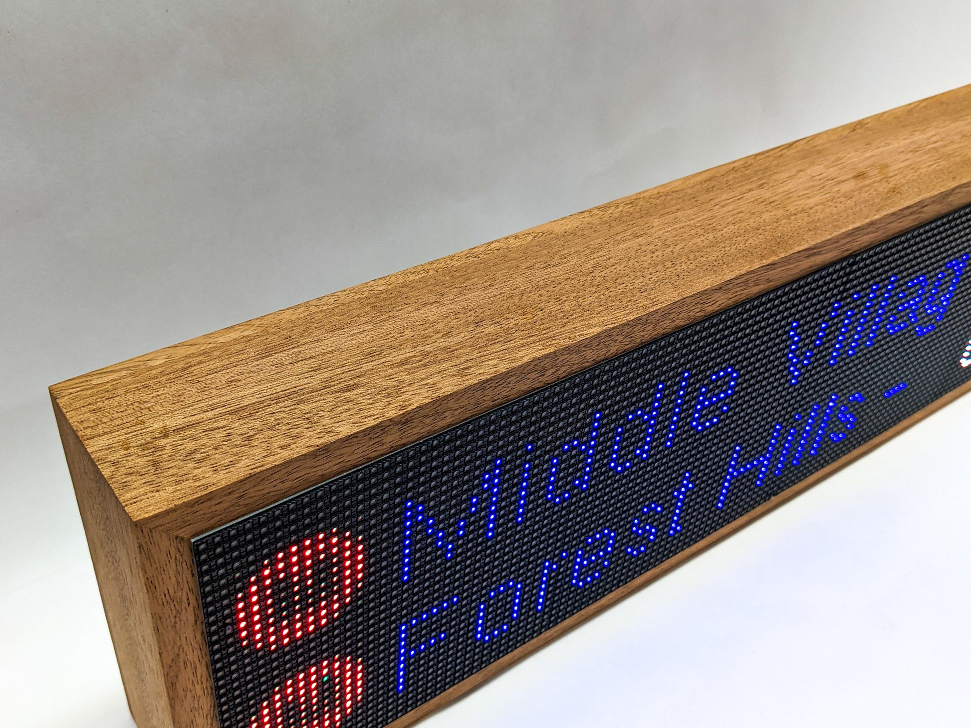 A top-view of the mahogany frame on the top of the NYC subway clock. THe mahogany is thick and angular with interesting darker streaks of wood grain. A black LED screen meets the wood with signs for the M train arrival times.