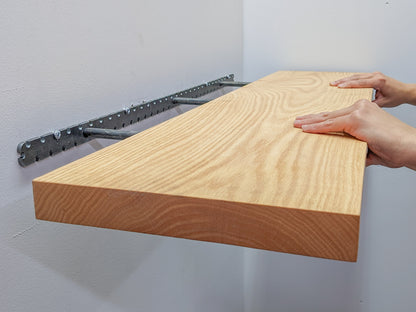 Two hands grip the top and bottom of the front piece of a long wooden oak shelf and easily install it onto the wall by sliding the shelf onto black brackets.