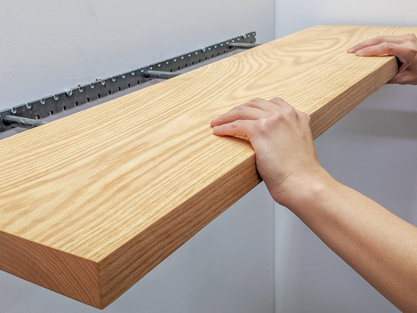 A caddycorner view of two hands gripping the top and bottom of the front section of a long oak standard shelf that is being pushed onto a black bracket.
