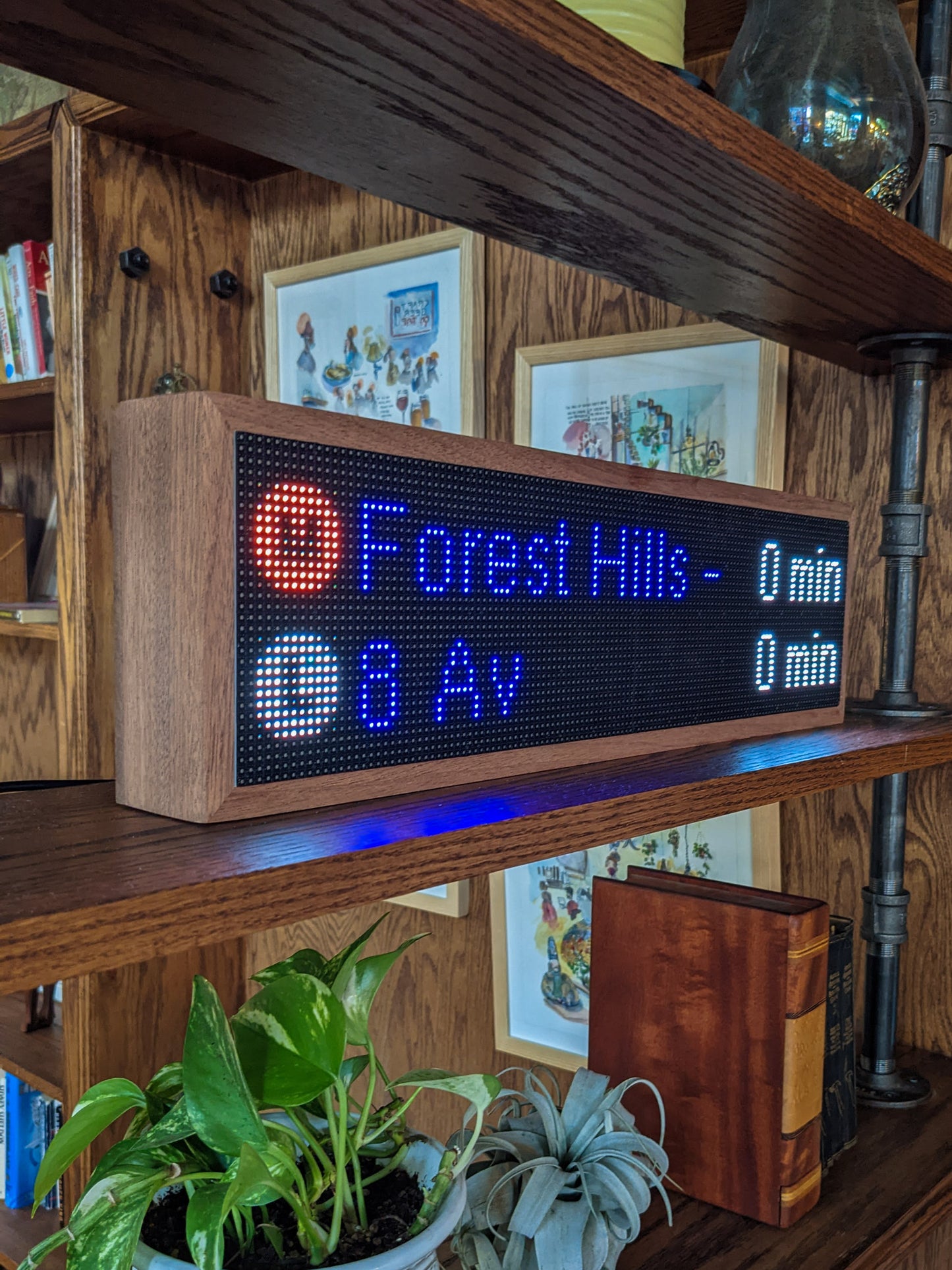 The NYC Realtime Subway Clock is angled on the middle shelf of a mahogany bookcase. The Mahogany frame holds a black LED screen that reads, "M Forest Hills -0 min" and below that "L 8 Av 0 Min" metal pipes hold the shelves up. A green potted plant, silver potted plant, and brown large leather book sit on the shelf below the subway clock.