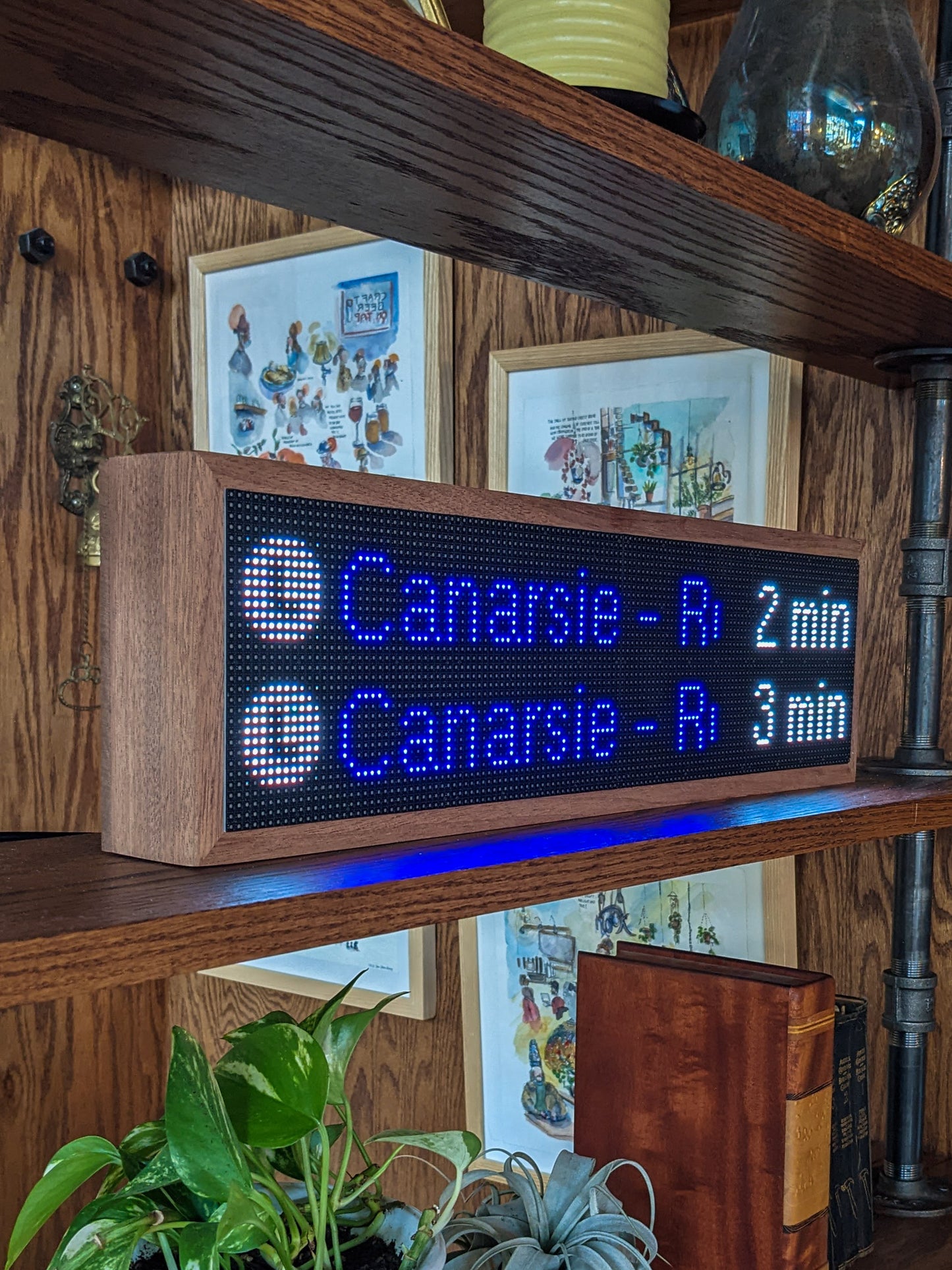 The NYC Realtime Subway Clock is angled on the middle shelf of a mahogany bookcase. The Mahogany frame holds a black LED screen that reads, "L Canarsie - R -2 min" and below that "L Canarsie -R  3 Min" metal pipes hold the shelves up. A green potted plant, a silver potted plant, and a brown large leather book sit on the shelf below the subway clock. In the back are four various illustrations that are framed and hang on the mahogany wood wall.