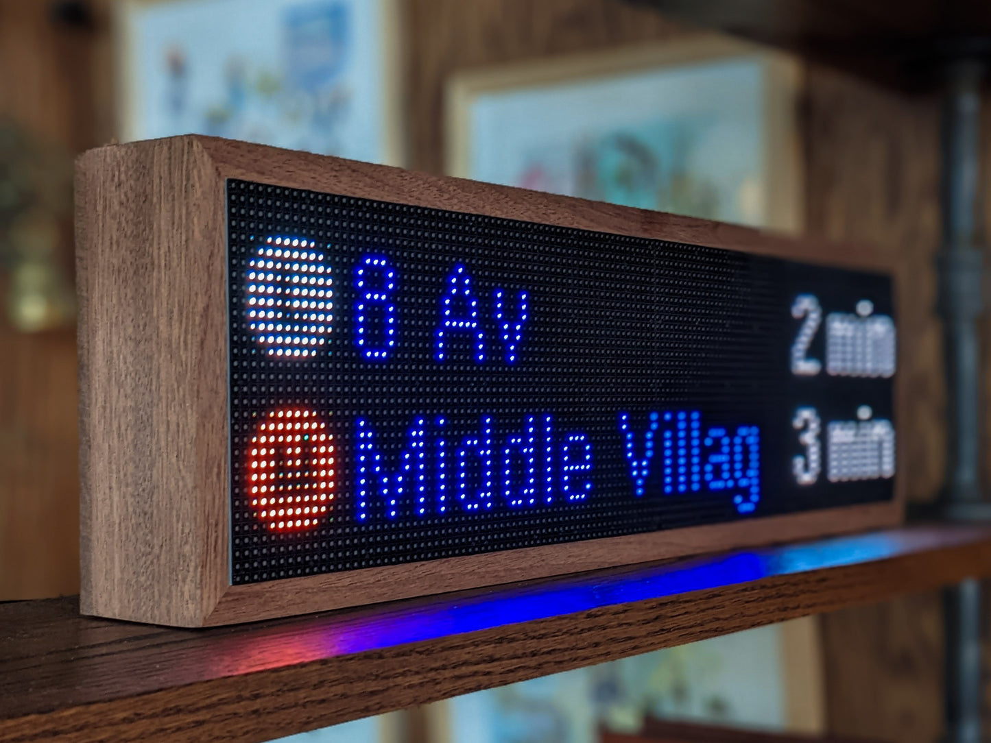 The NYC Realtime Subway Clock is angled on a mahogany shelf. Framed in mahogany the black LED screen features a round white circle with the letter L in it next to the words, 8 Av next to "2 min" on the left side. Directly below, a round orange circle has the letter M in it next to blue "Middle Village" which is next to a white "3 min".
