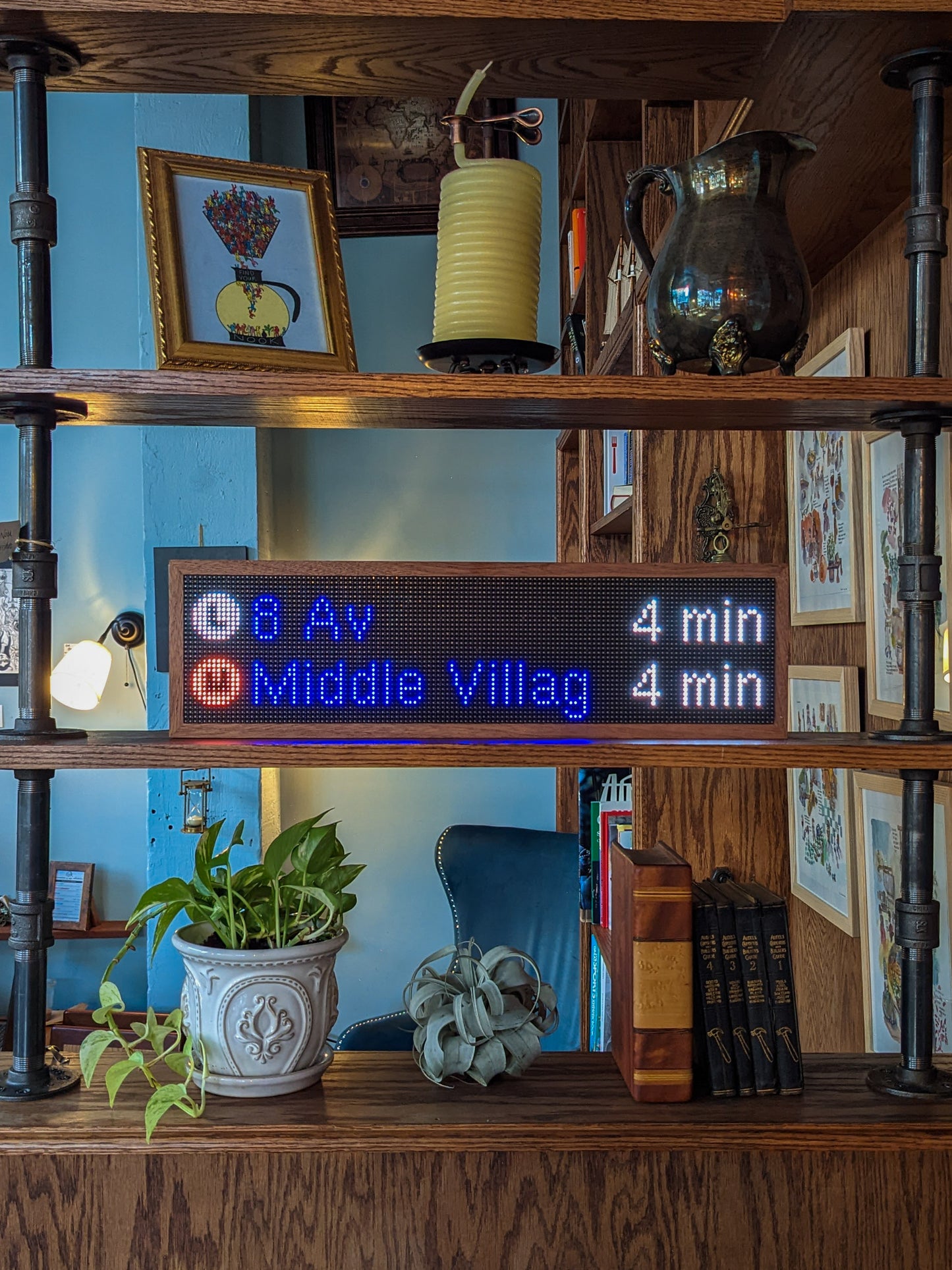 Three shelves of a mahogany bookcase are on display. The top shelf holds an illustration of a coffee carafe, wick candle, and antique pitcher, the middle shelf holds the subway clock with mahogany frame. The subway clock reads, "L 8 Av 4 min" and below that, "M Middle Villag 4 Min", on the bottom shelf a green plant sits in an intircate white planter next to a silver air plant, a large brown book, and four black books. The wall behind it is a light blue.