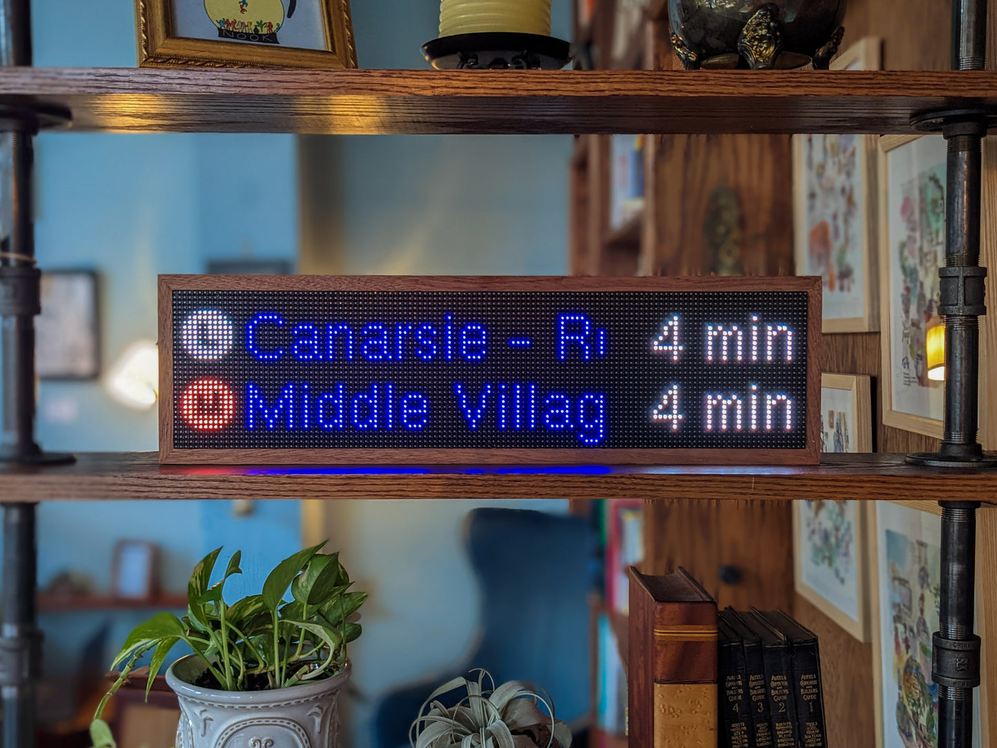 The MTA Realtime Subway Clock sits on the middle shelf of a mahogany bookcase. Mahogany frames a black-led screen that reads, "L Canarsie - R 4 min" and below that "M Middle Villag 4 Min". The shelf above holds a gold-framed picture and candle that are cut-off from view. The shelf below holds a potted green plant, a silver plant, a thick brown leather book and 4 black leather books with gold embossing.