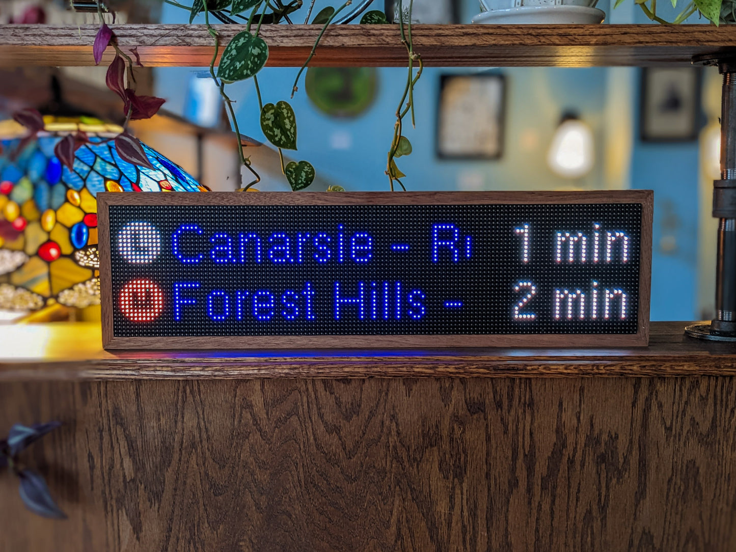 An NYC Subway Clock sits on a mahogany counter. A mahogany frame holds a black LED screen which reads, "L Canarsie- R 1 min" and directly below that on the screen "M Forest Hills- 2 min". A mahogany shelf sits above the clock and various plant vines drape artistically over the clock. A tiffany style lap is in the left back.