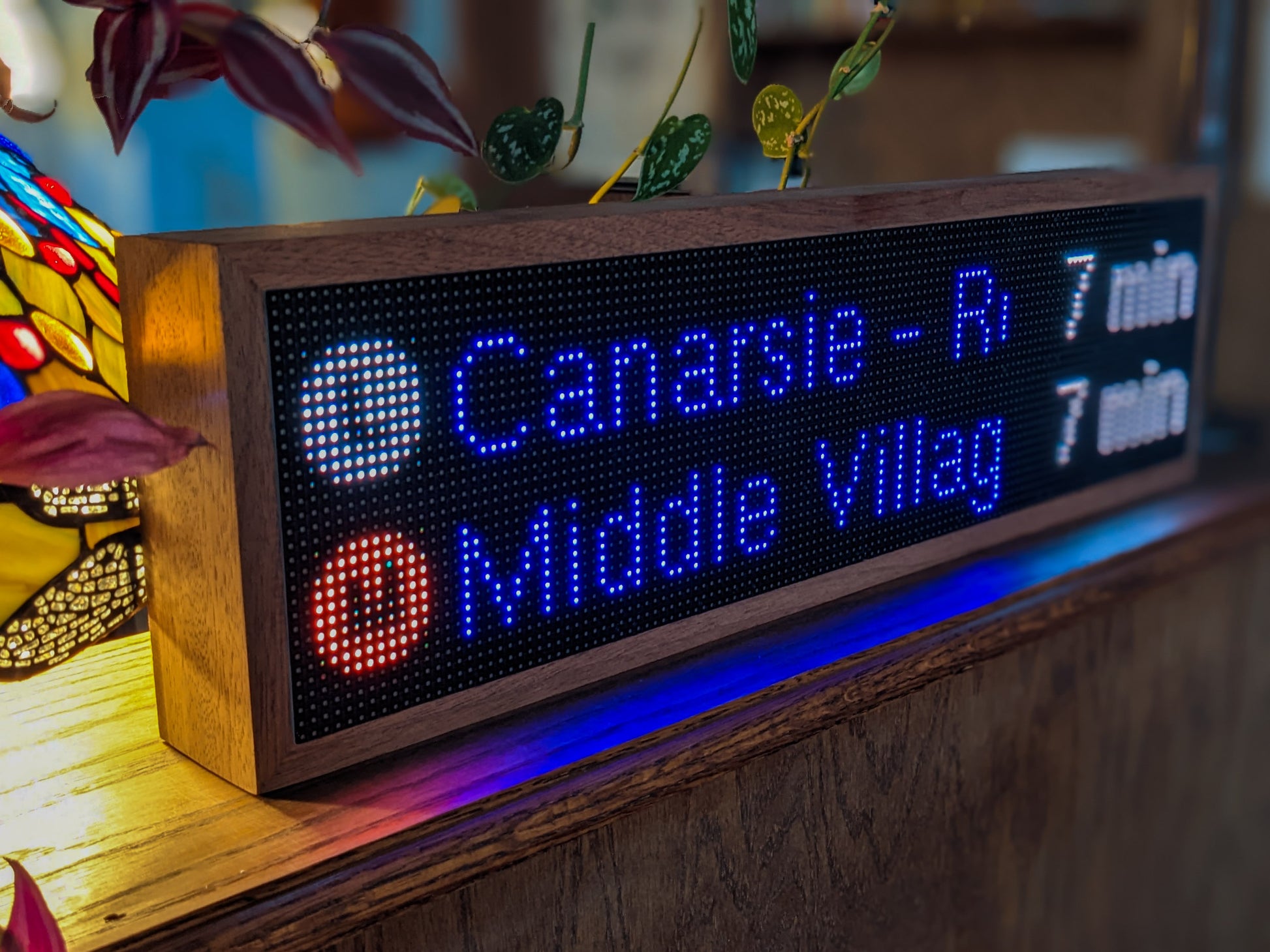 The NYC Realtime Subway Clock is angled on a mahogany shelf. The variagated leaves and vines of plants drape near it. The Mahogany frame hold a black LED screen that reads, "L Canarsie -R 7 min" and "M Middle Villag 7 min" A tiffany style lamp glows warmly to the left.