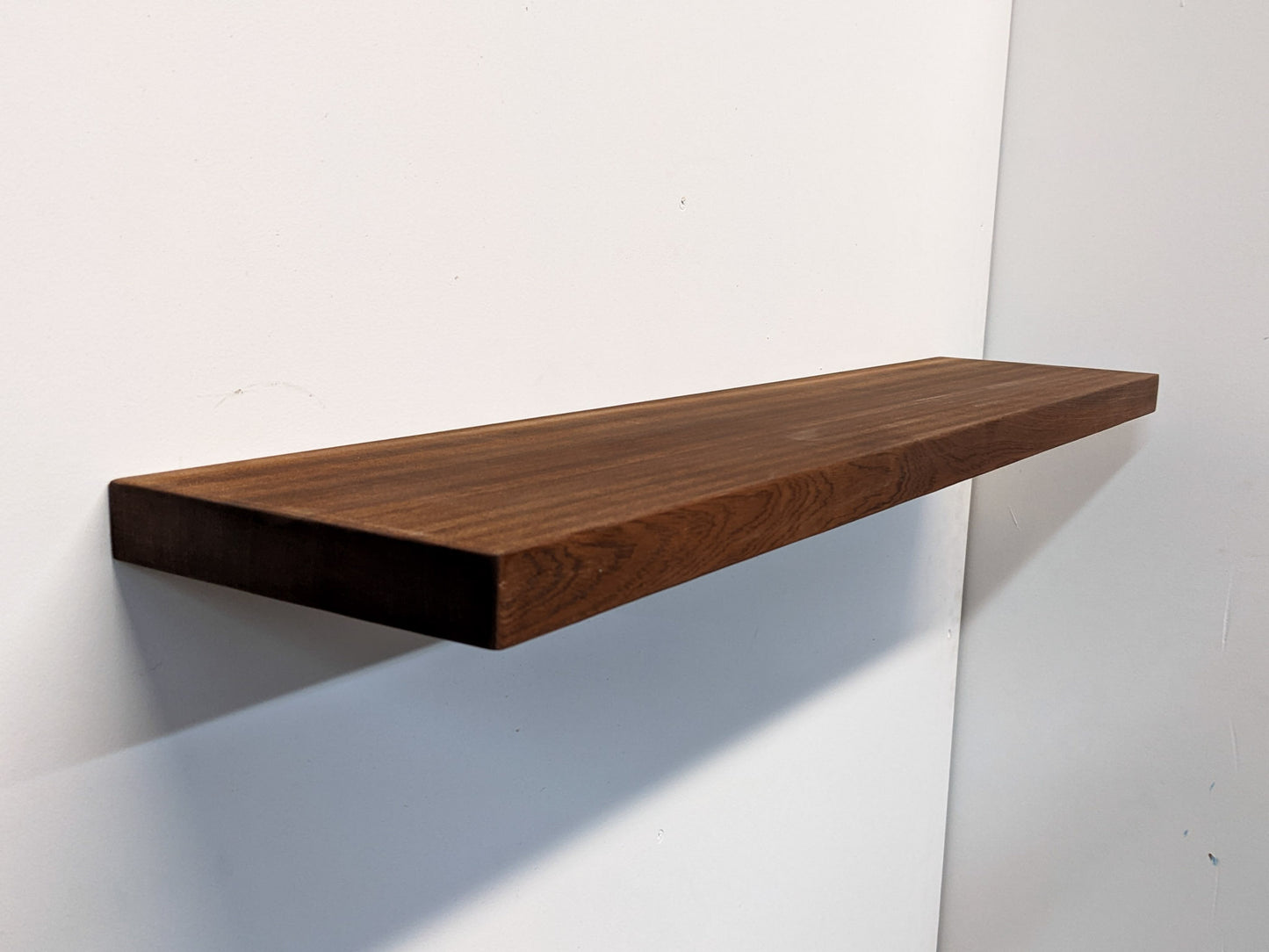 A long floating shelf floats on a wall. The brown mahogany wood displays the natural grain of the heavy duty shelf is highlighted. The shelf casts a shadow on the wall.