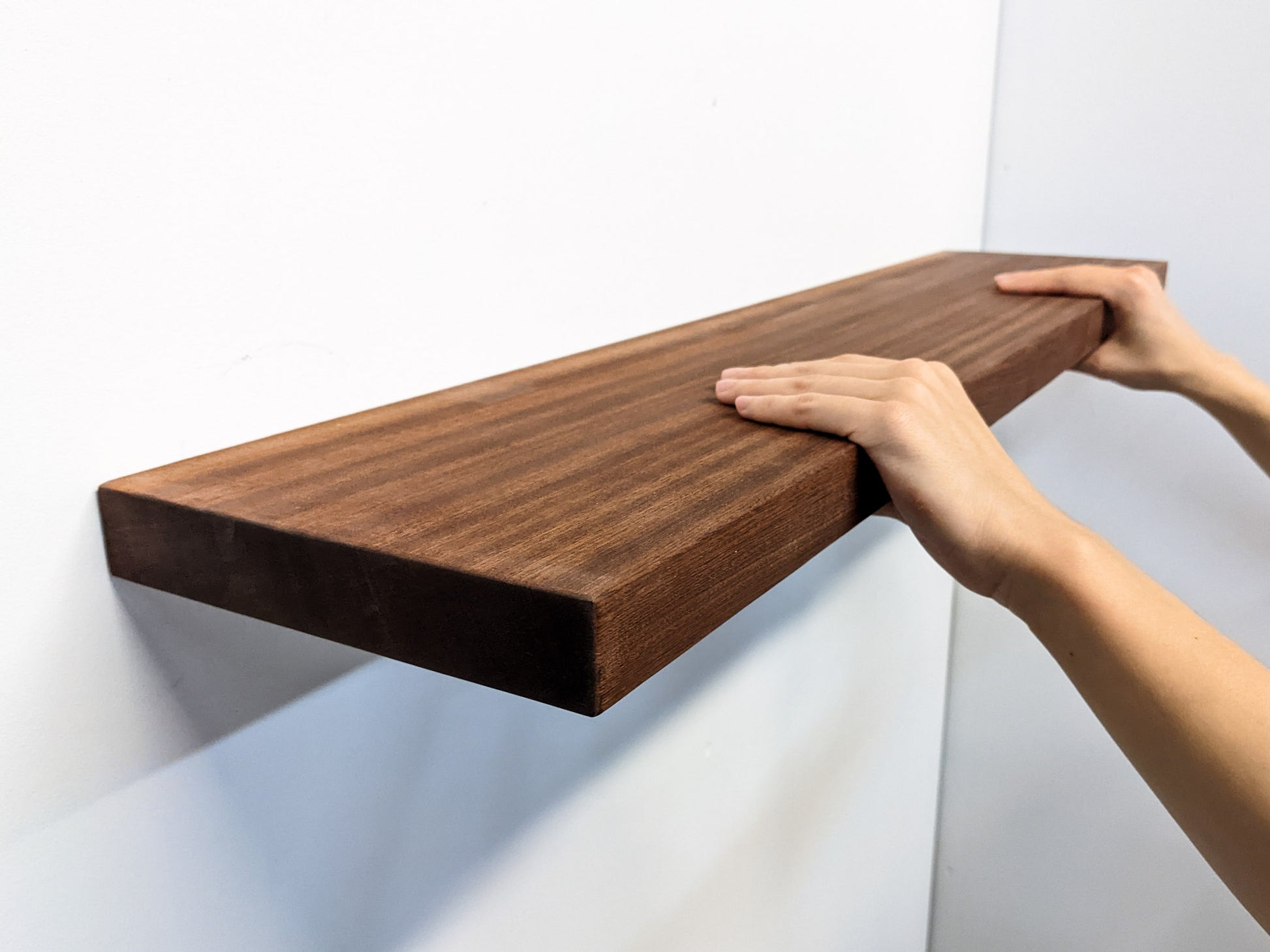 An extra-long, heavy-duty mahogany floating shelf is sucessfully installed and sits securely on a wall. Two hands grip the top and bottom of the front-facing side.