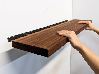 Two hands grip the front of a long wooden mahogany shelf and easily install it on the wall by sliding it onto black brackets. 